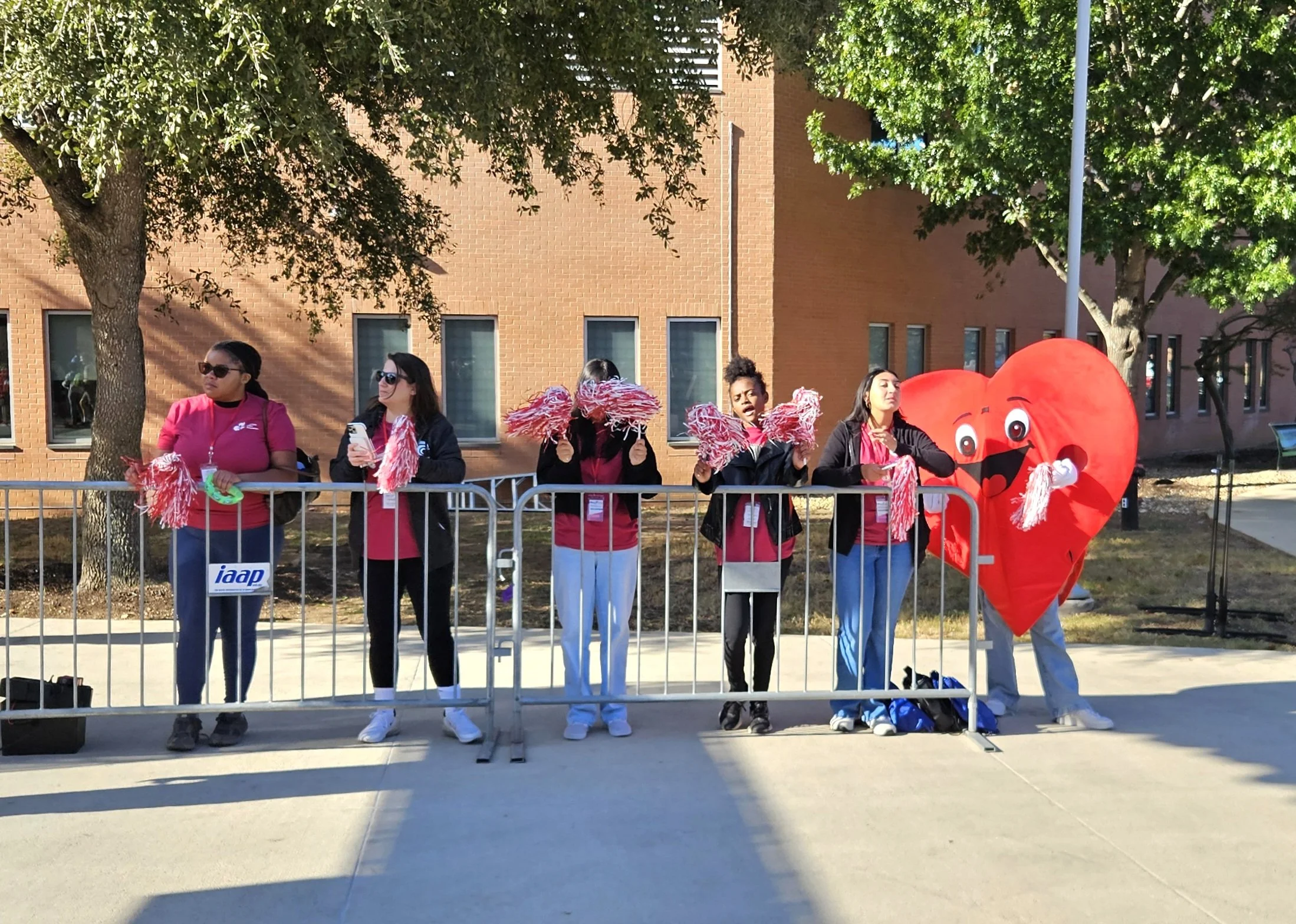 Group of women and children at a heart-themed event outdoors, some holding pink pom-poms, with a large inflatable heart costume.