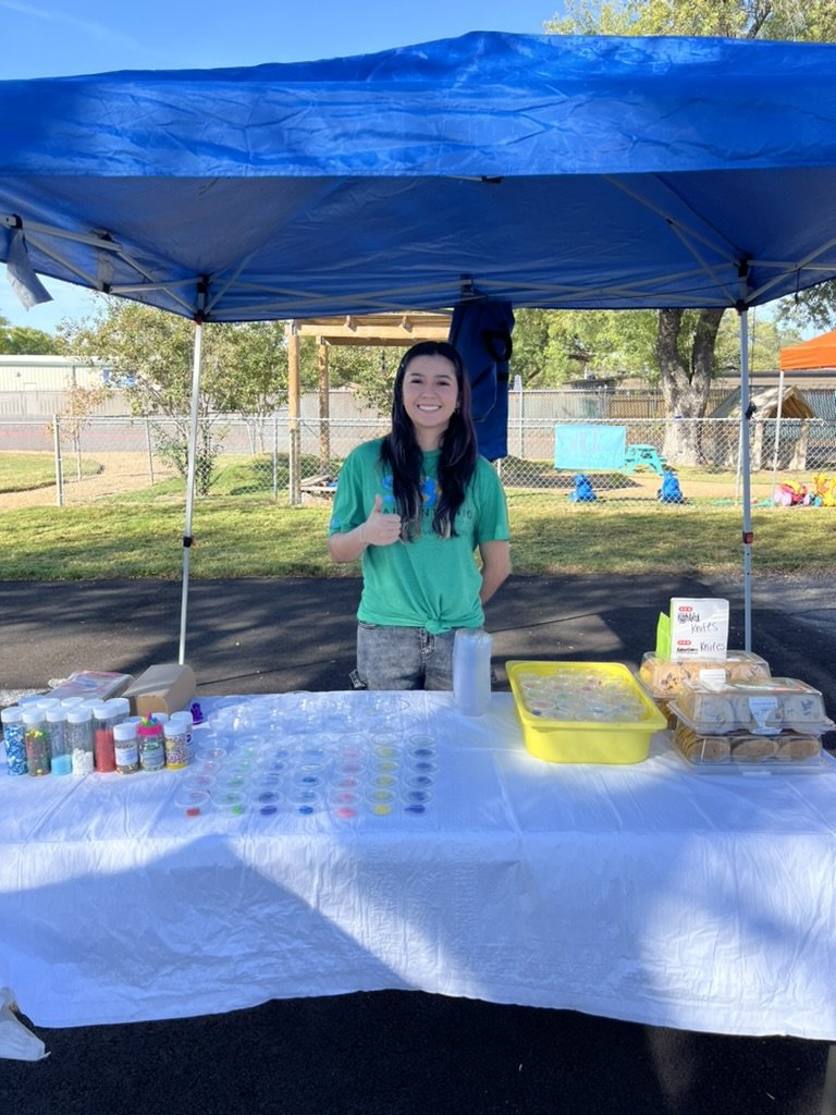 Young woman standing behind a table at an outdoor event, under a blue canopy, with various small containers and baked goods on display.