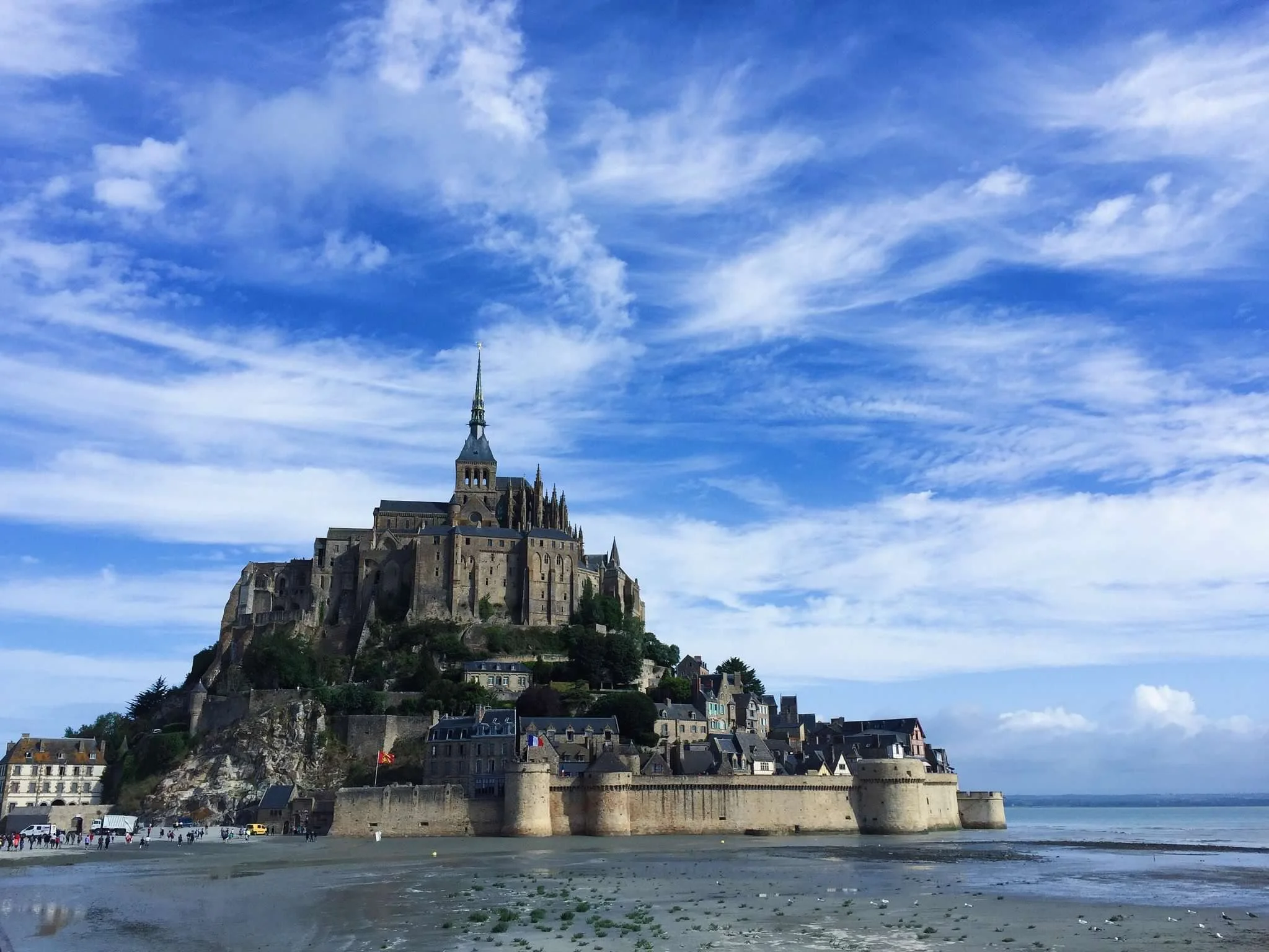 Mont Saint-Michel, a historic island commune with a medieval monastery and fortress, surrounded by water and tied to the mainland by a causeway during low tide, under a blue sky with scattered clouds.