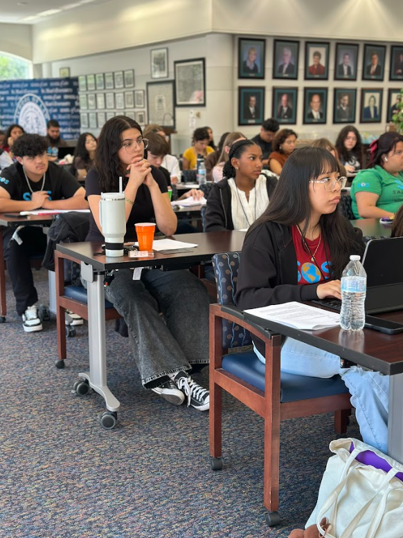 A diverse group of students attending the St. Mary's University Law School, sitting at desks with notebooks, drinks, and electronic devices, listening attentively.