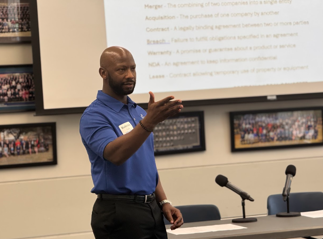 A man in a blue shirt standing in front of a large screen with a presentation slide, gesturing with his right hand during a speech or lecture in a conference room.