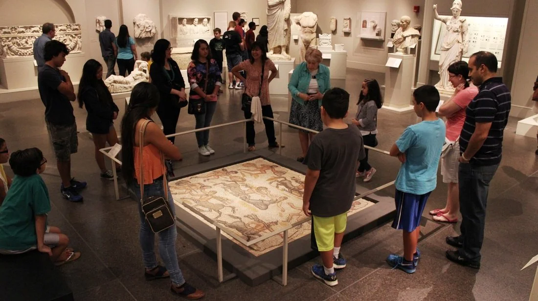 Group of children and adults observing an ancient mosaic in a museum, surrounded by classical sculptures and artifacts.