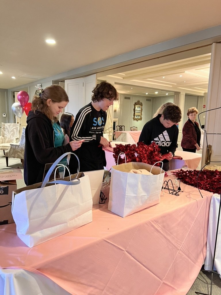 People wrapping red gift decorations at a table with pink tablecloths, with gift bags and balloons in the background.