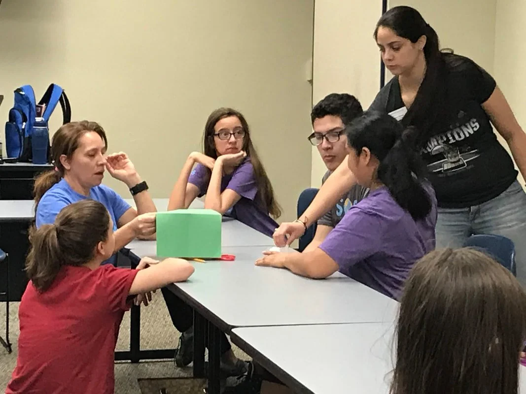 A group of kids sitting around a table with one girl standing and demonstrating something with her hands, while adults and children observe, in a classroom or meeting room.