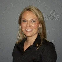 A woman with blonde hair smiling, wearing a black top, against a gray background.