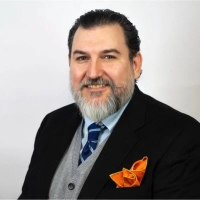 A man with dark hair and a beard smiling, wearing a black suit jacket, light blue shirt, striped tie, gray vest, and an orange pocket square, standing against a plain light background.