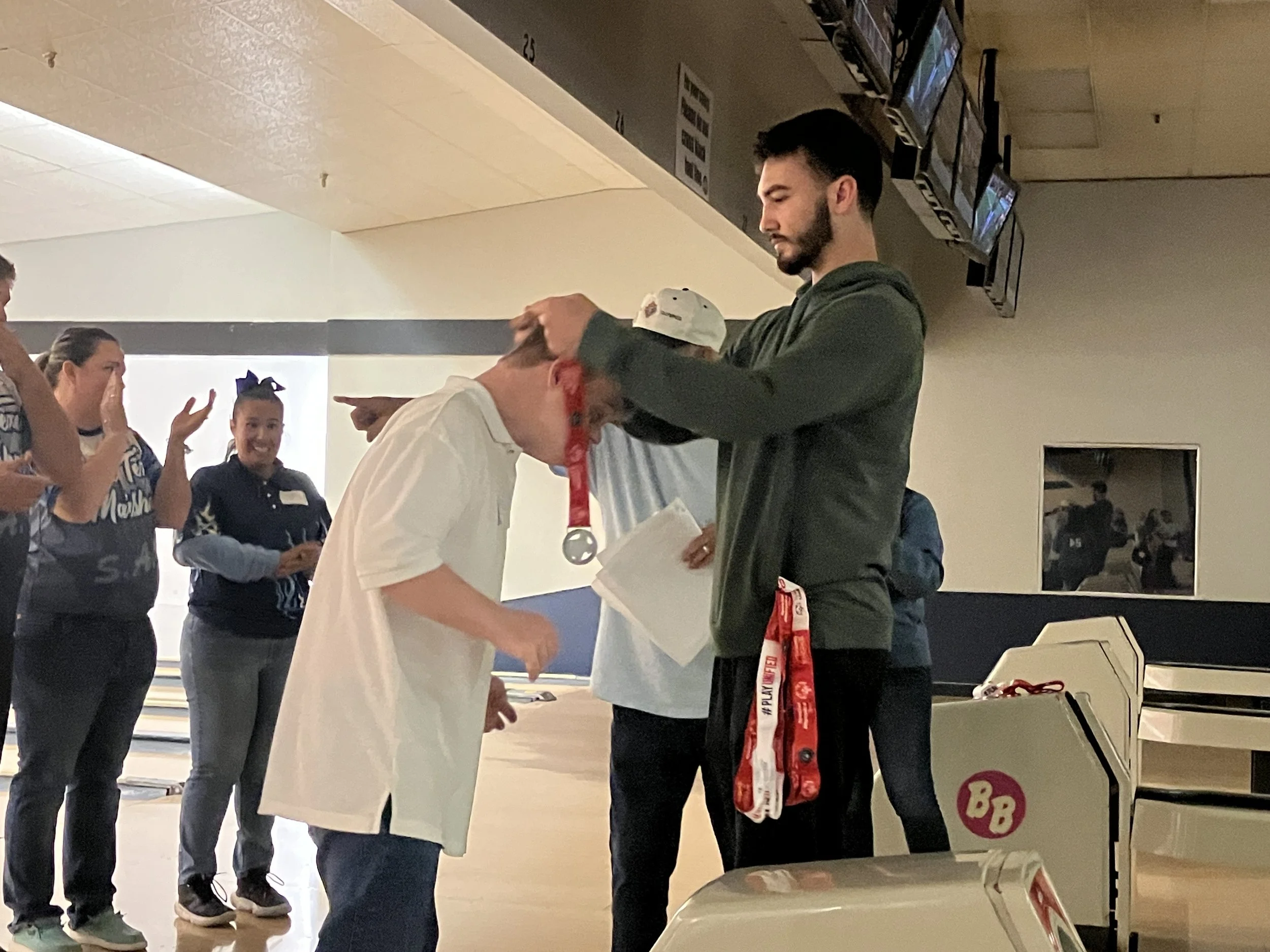 A man is placing a medal around the neck of a young person at a bowling alley during an award ceremony, with other people clapping and smiling in the background.