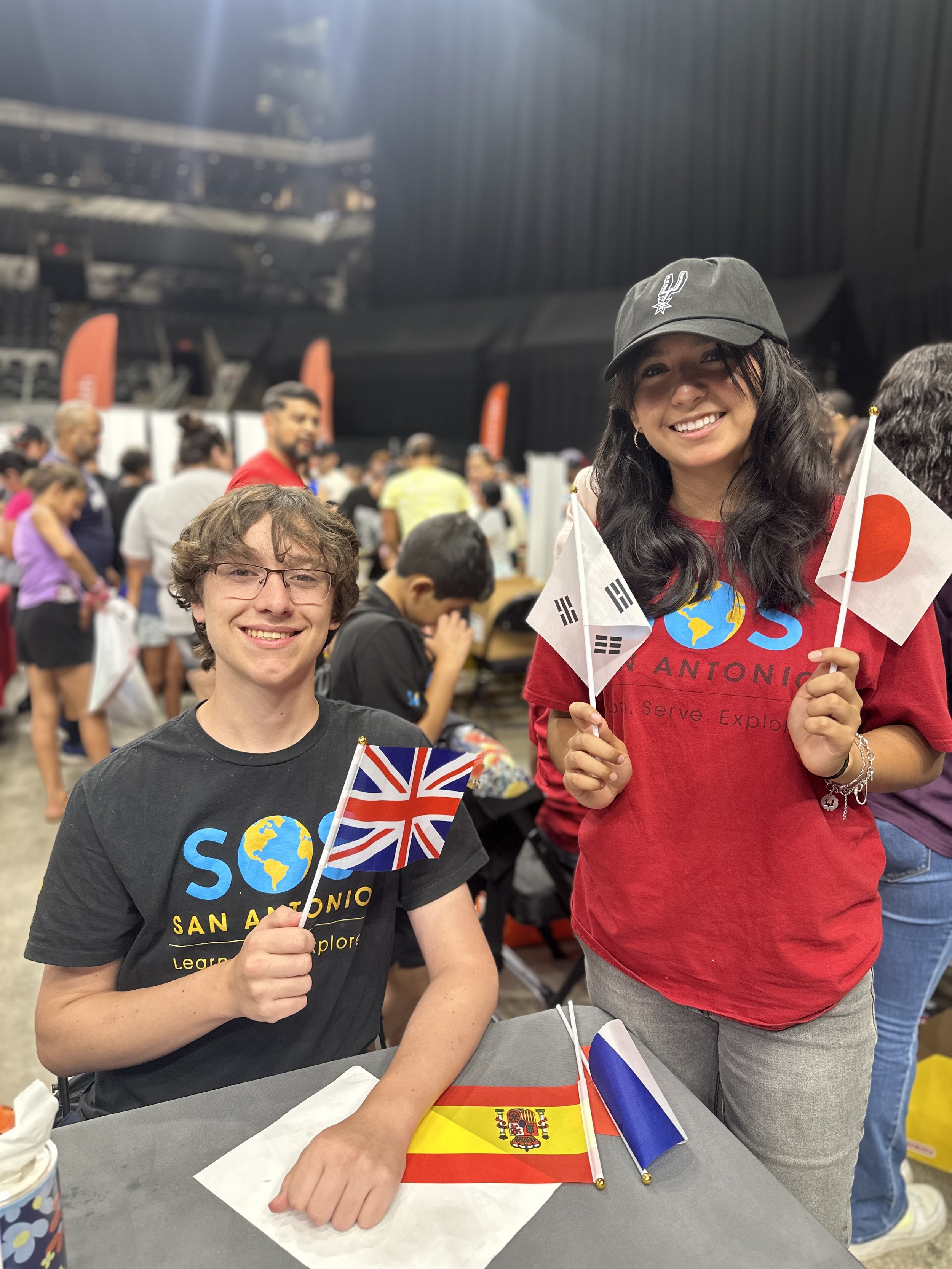 Two smiling young women holding international flags at a crowded event. One woman is seated at a table with a Spanish flag, and the other is standing with Japan and South Korea flags. Both are wearing T-shirts with a globe and the word 'SAN ANTONIO'.