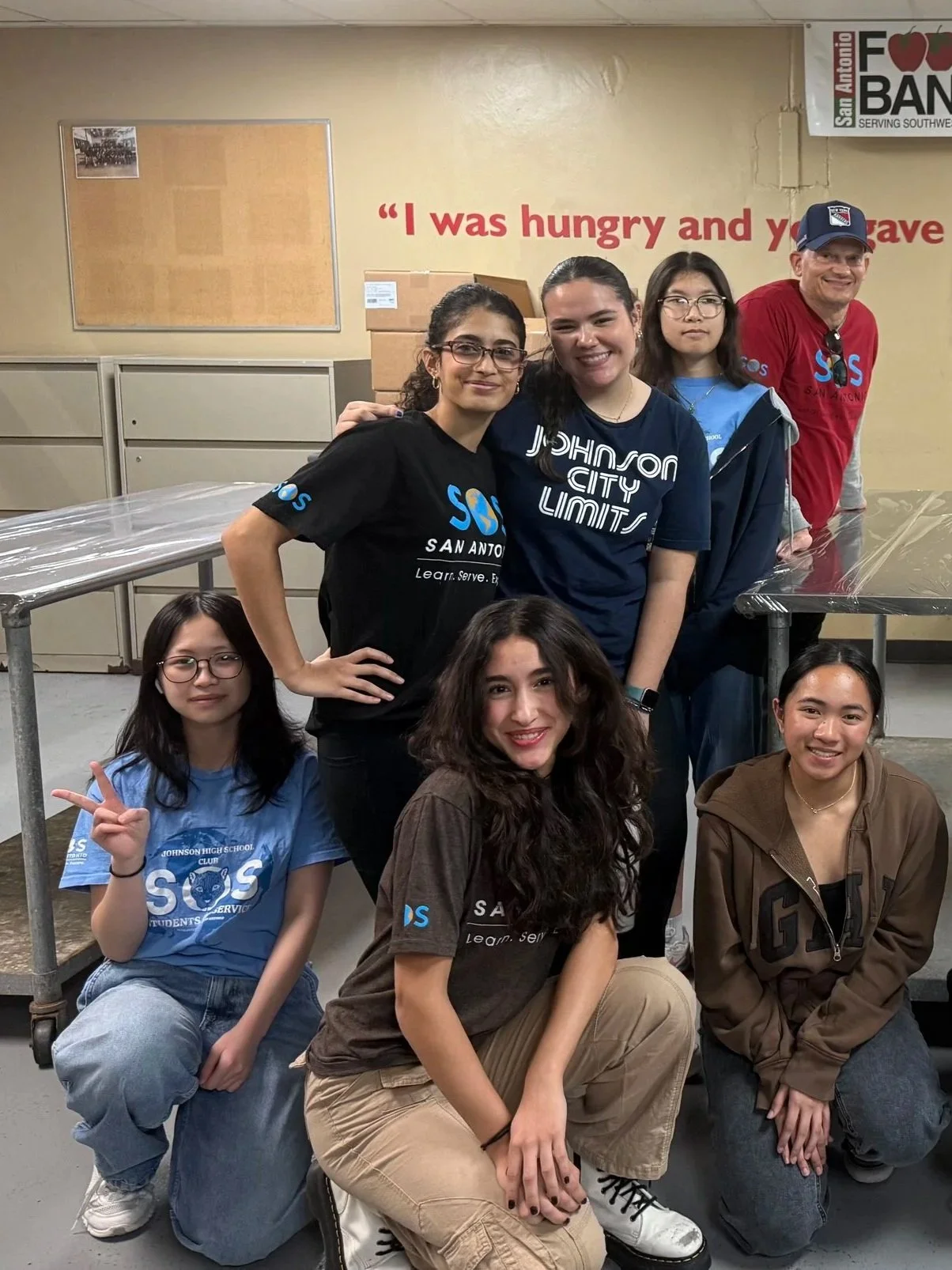 Group of young people posing together at a food bank, with some making peace signs. They are wearing casual clothes and are in a room with a Food Bank sign and boxes in the background.
