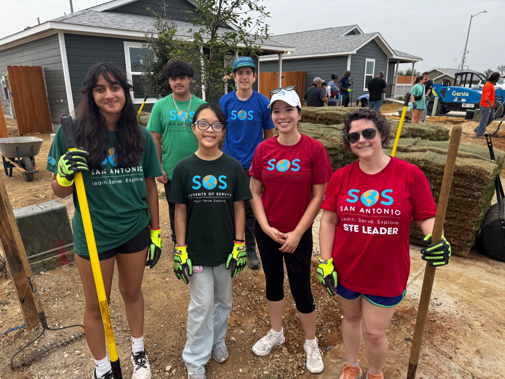 Group of six young volunteers at a community landscaping project, holding tools, with a house and other people working in the background.