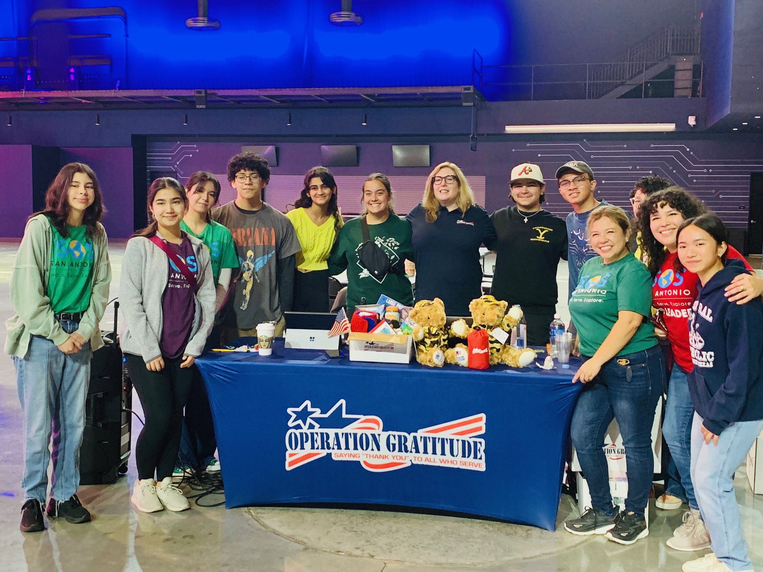 Group of young people and adults standing behind a table with a blue tablecloth that reads 'Operation Gratitude,' at an indoor event. The table has various items on it, including small stuffed animals and U.S. flags. Everyone is smiling and posing fo