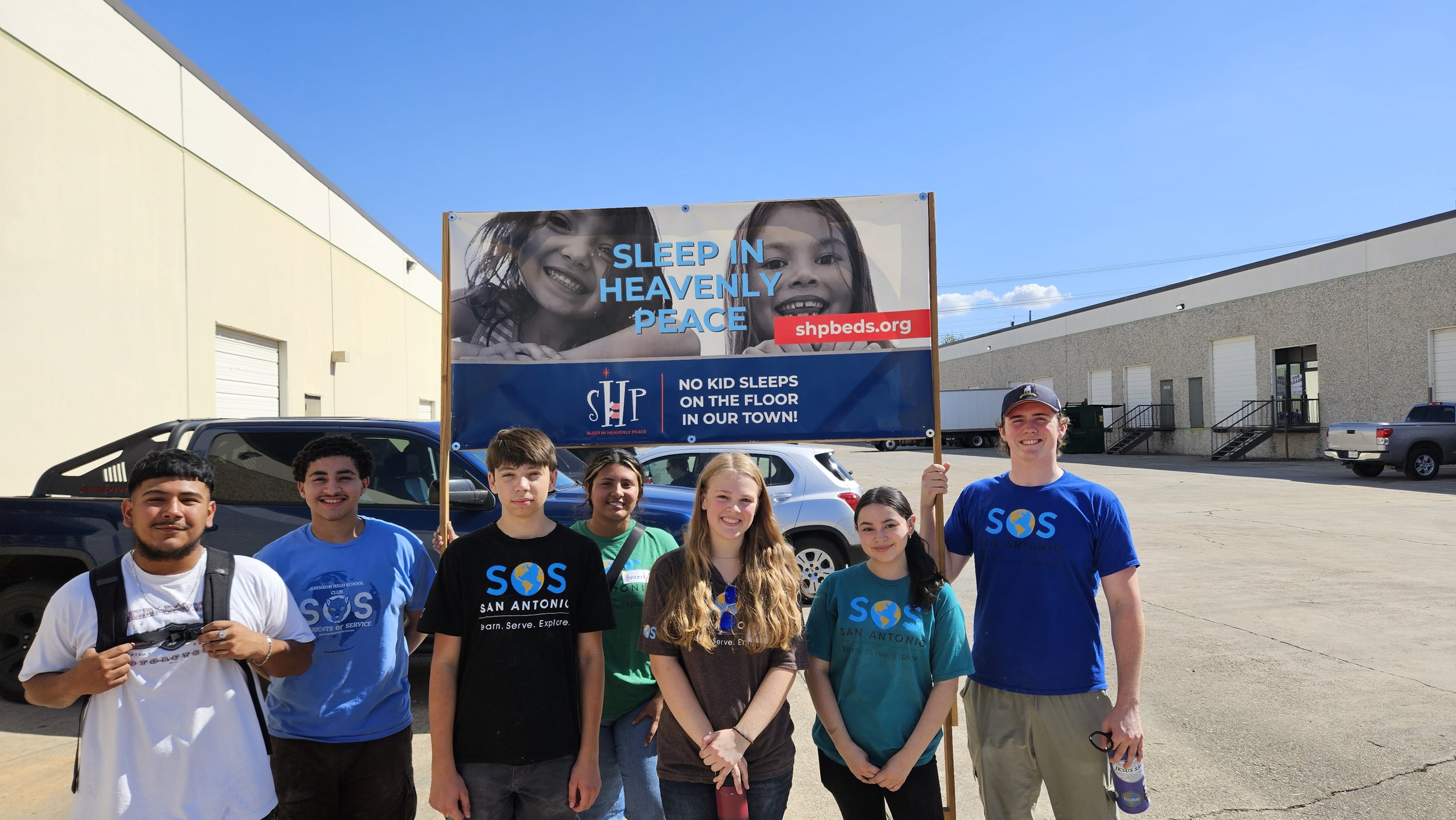 Group of seven teenagers standing outdoors in front of a large sign that reads "Sleep in Heavenly Peace" and "No kid sleeps on the floor in our town," with a logo and website address. They are smiling, some wearing shirts with "SOS San Antonio" print