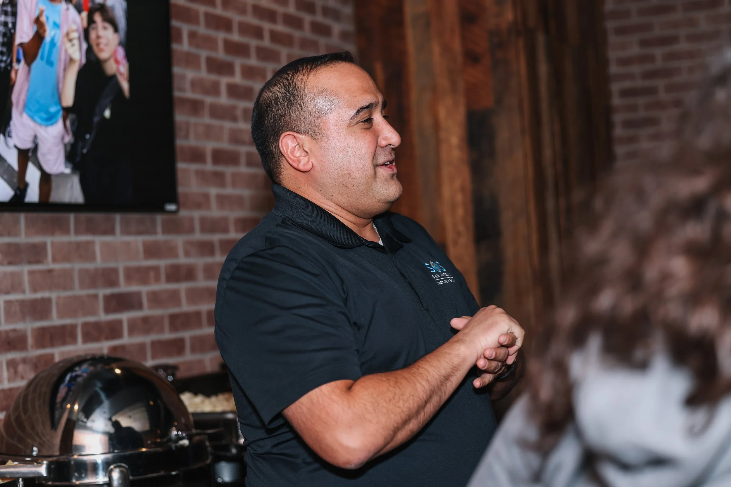 A man in a black polo shirt with a logo stands with his hands clasped in front of a brick wall and a flat screen TV. There is a shiny metal serving dish in the foreground.