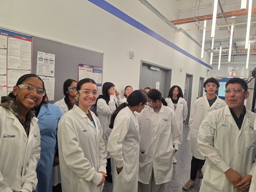 Group of students in white lab coats and safety glasses standing in a hallway with informational posters on the wall.