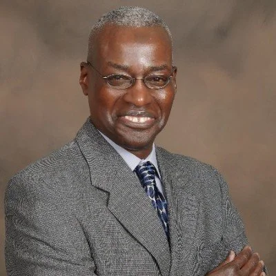 A professional man with gray hair and glasses, wearing a gray suit and a patterned tie, smiling with arms crossed against a neutral background.
