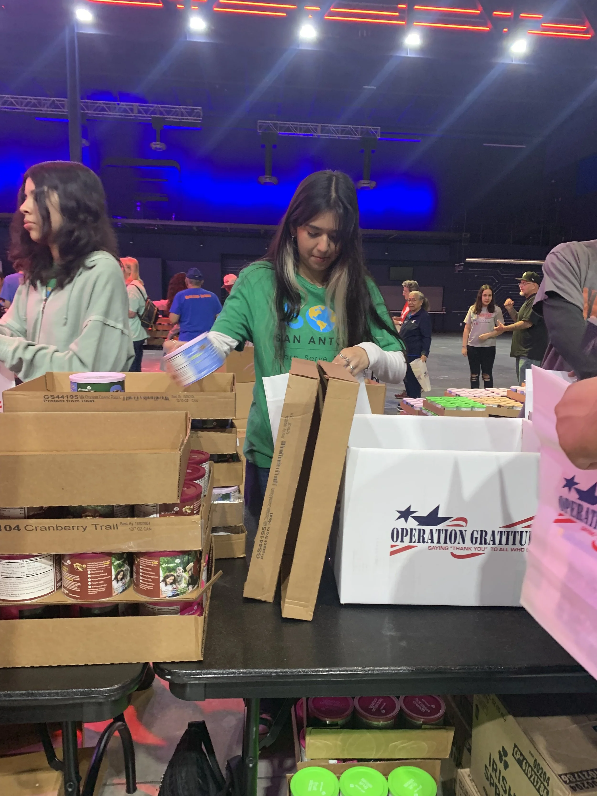 People volunteering at a charity event, organizing food and supplies at a table labeled 'Operation Gratitude.' The background features a large dark indoor space with blue and red lighting.