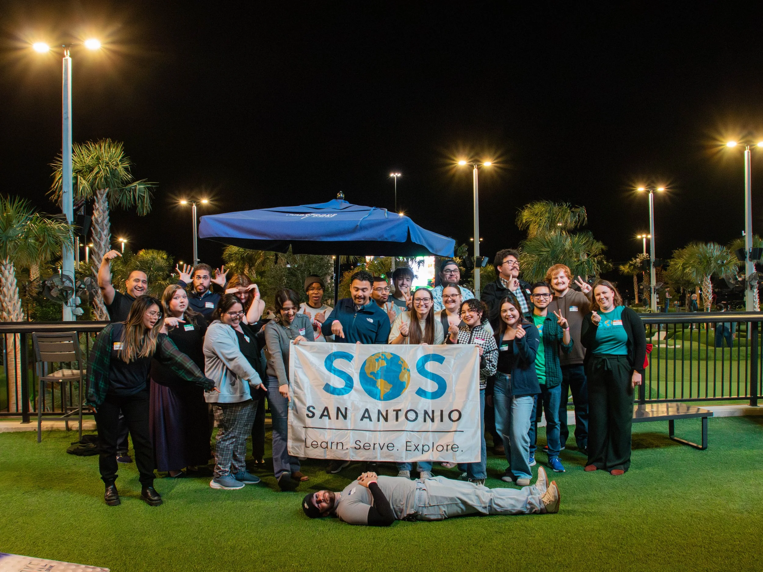 Group of people holding a SOS San Antonio banner at night outdoors, smiling and posing for a photo, with palm trees and streetlights in the background.