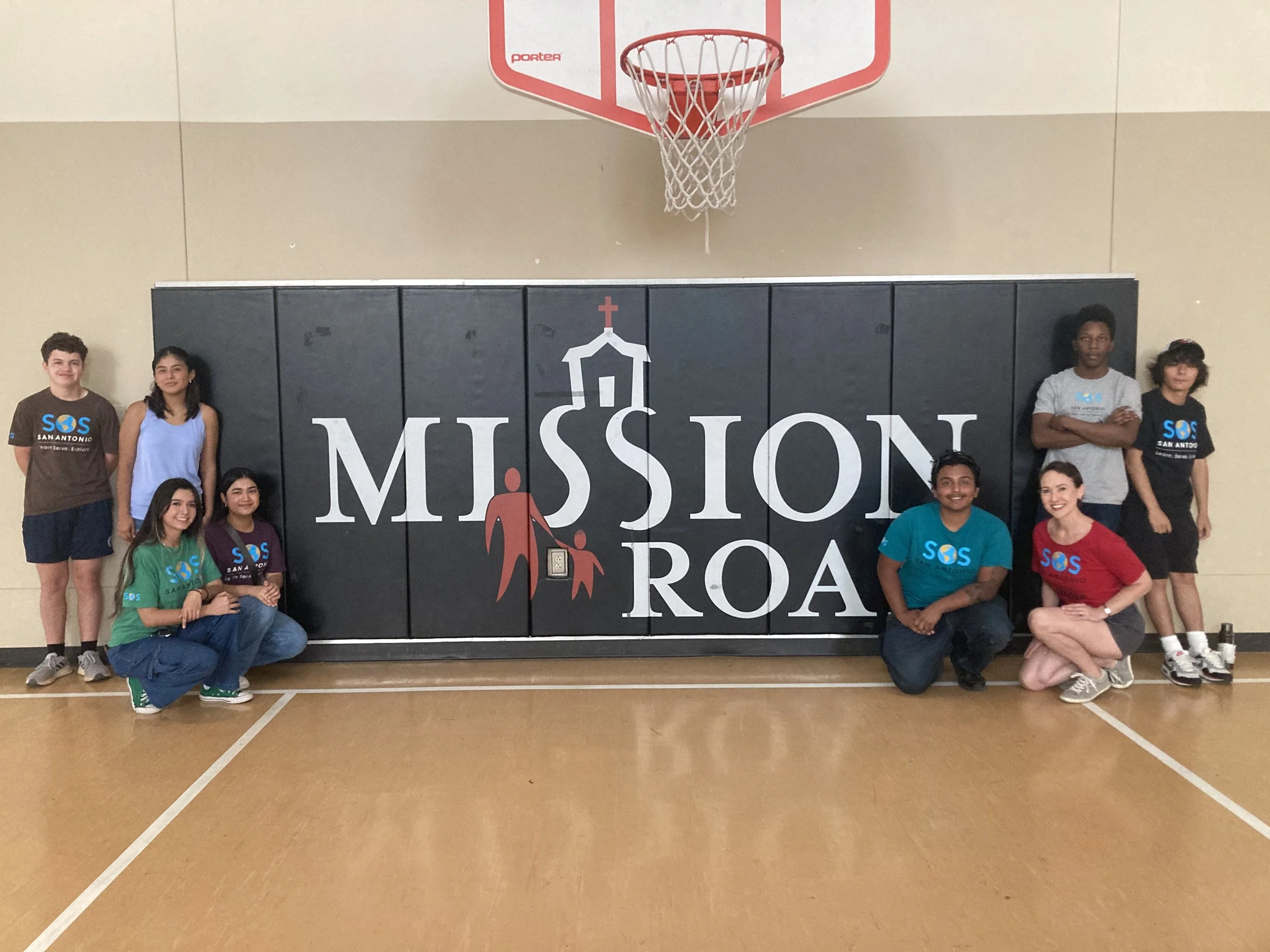 Group of nine young people standing and kneeling in front of a black wall with a Mission Road logo, inside a gymnasium with a basketball hoop overhead.