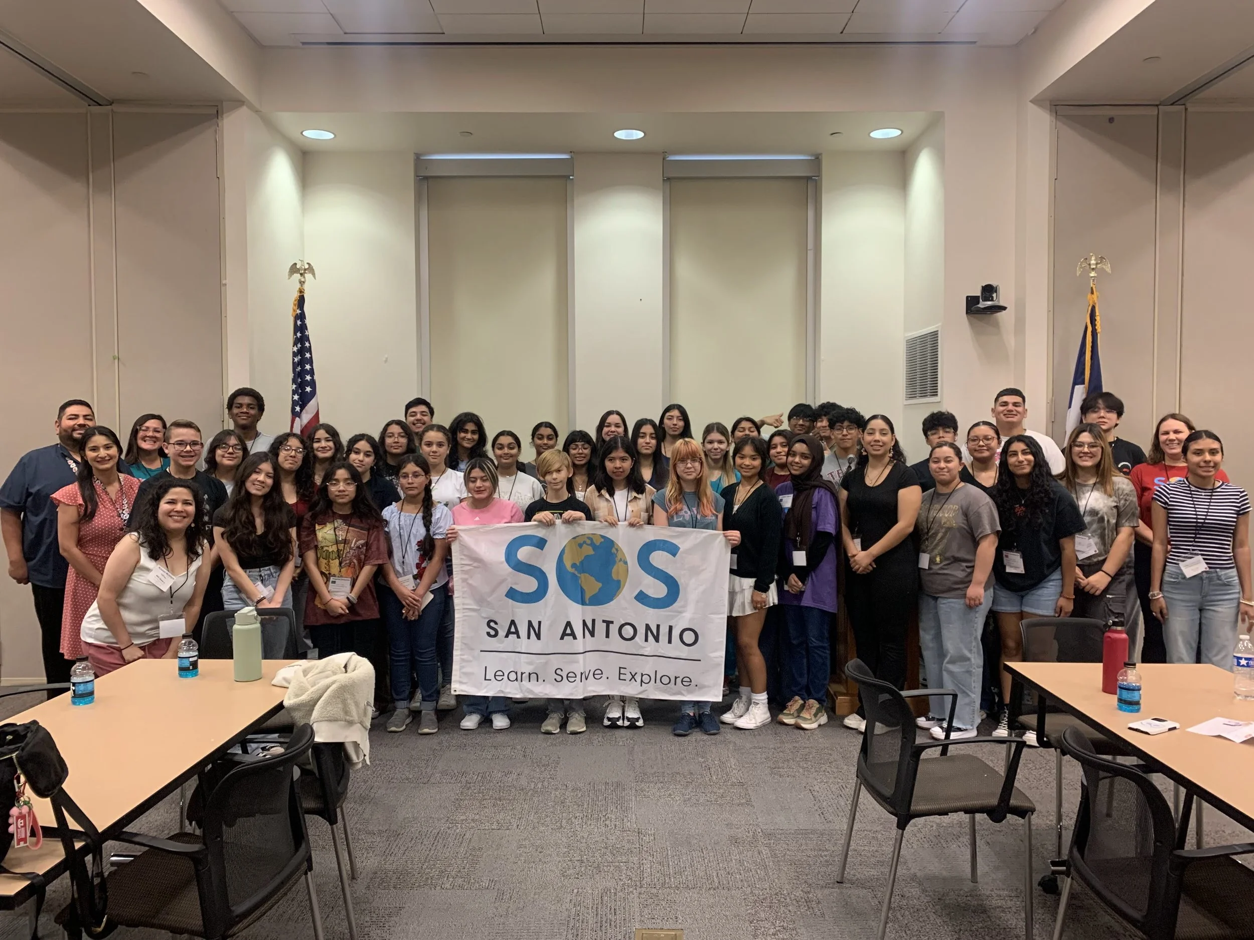 Group photo of diverse students and adults holding a large SOS San Antonio banner with the tagline "Learn. Serve. Explore." in a conference room with American and Texas flags, tables, chairs, and water bottles.