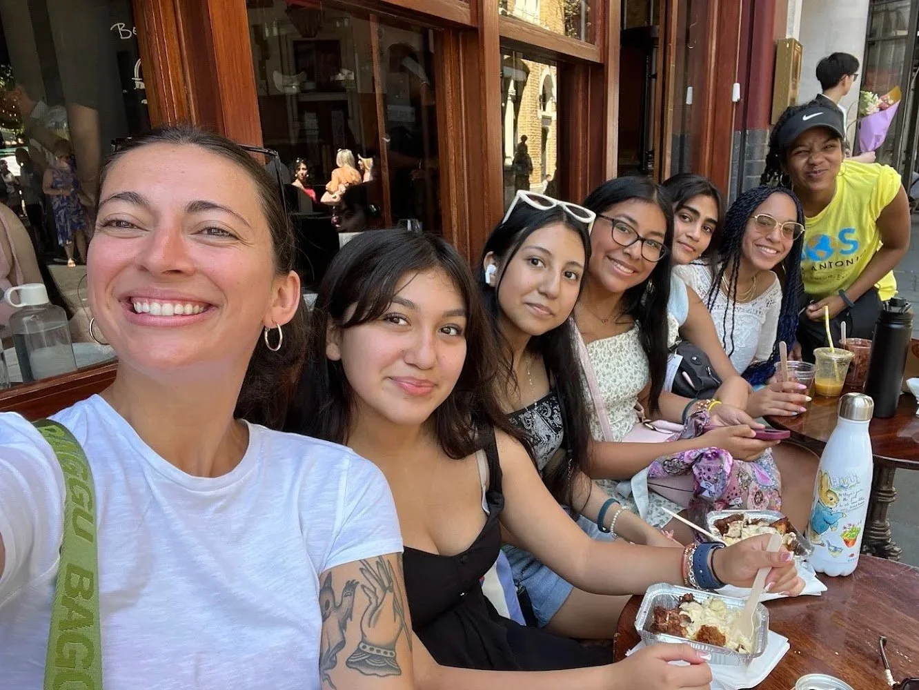 A group of seven women sitting at a wooden outdoor cafe table, smiling and enjoying desserts and drinks, with a woman taking a selfie.
