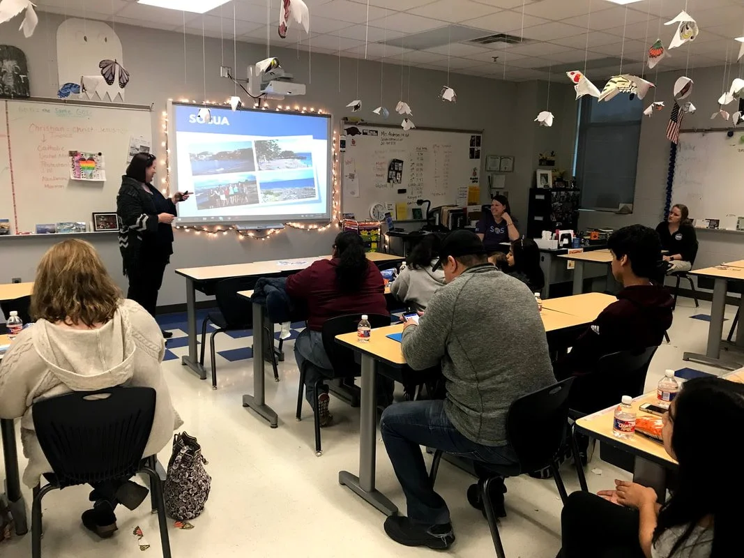 A classroom with students sitting at desks, listening to a woman giving a presentation in front of a screen displaying images. The classroom is decorated with hanging paper butterflies and paper boats.