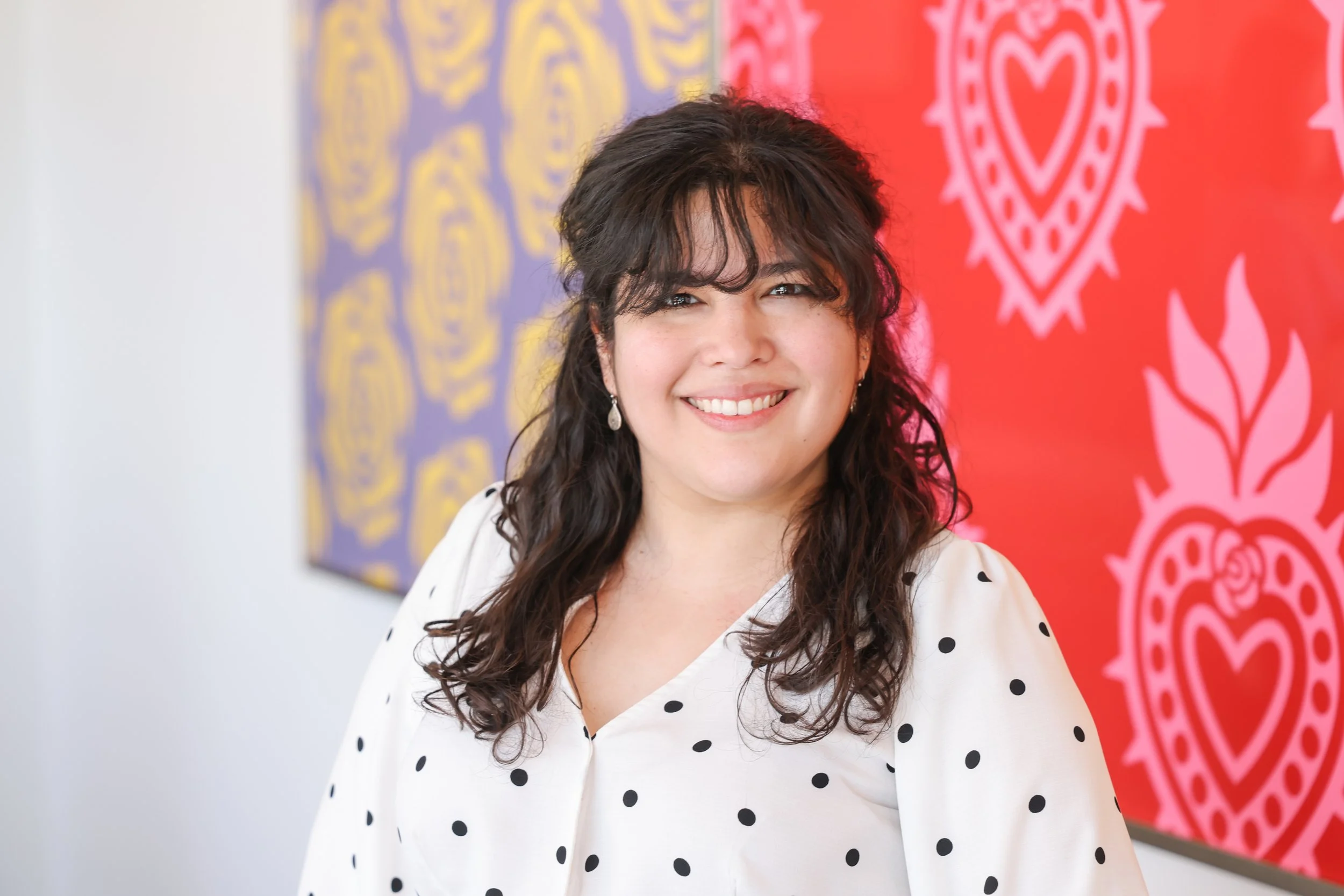 A woman with dark curly hair and earrings smiling, wearing a white blouse with black polka dots, standing in front of colorful, patterned wall art with red and purple backgrounds featuring heart and floral designs.