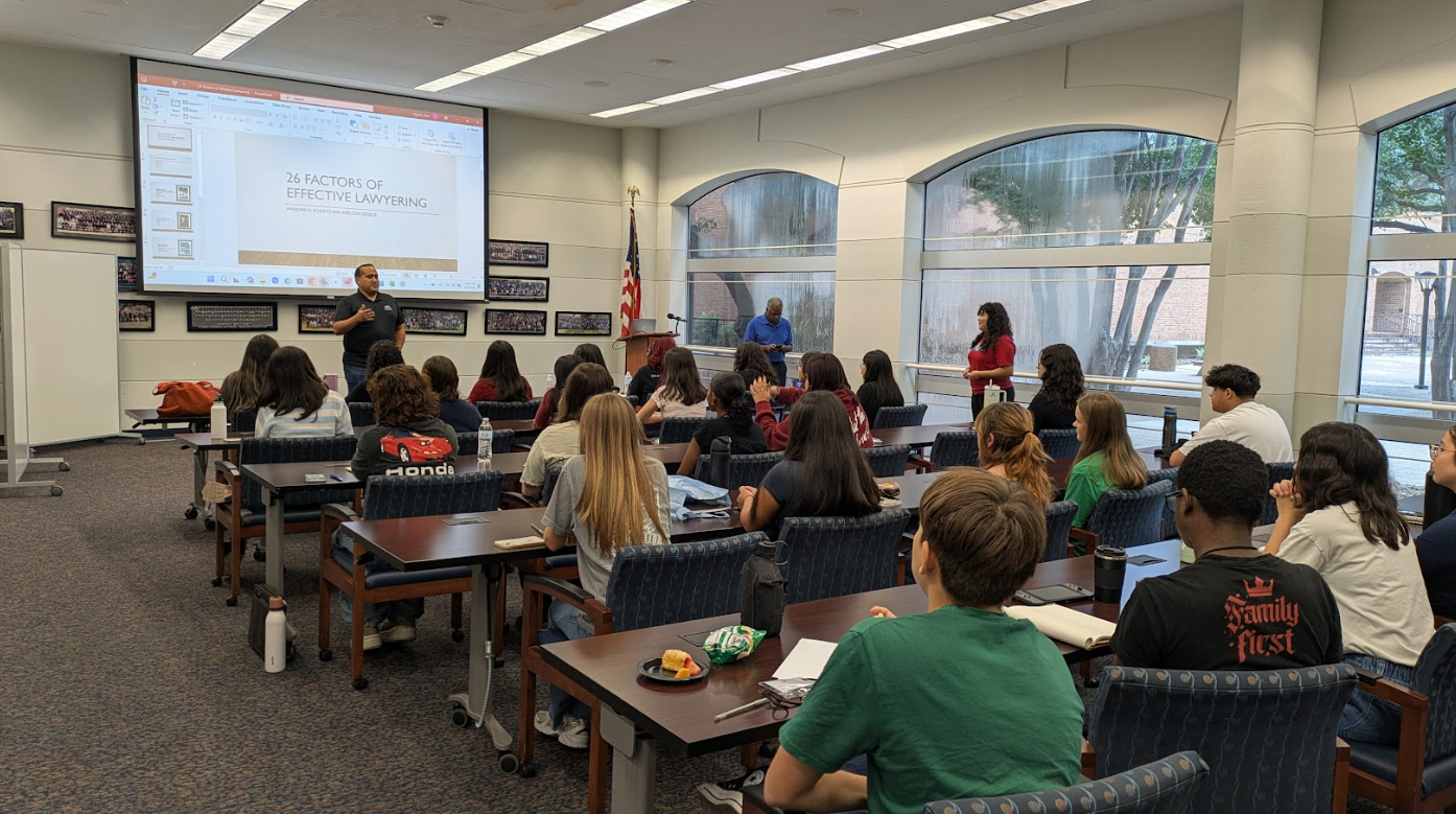 A seminar room with students seated at tables, listening to a presentation about factors of effective lawyering. A man is speaking at the front near a large screen, and a woman is standing to the side.