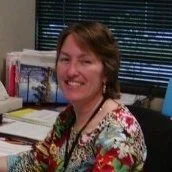 A woman smiling at a desk in an office, wearing a floral blouse and glasses.