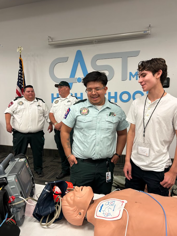 Group of four young individuals in a medical training room, with a CPR training mannequin on the table, and a background sign that reads 'CAST MH High School.'