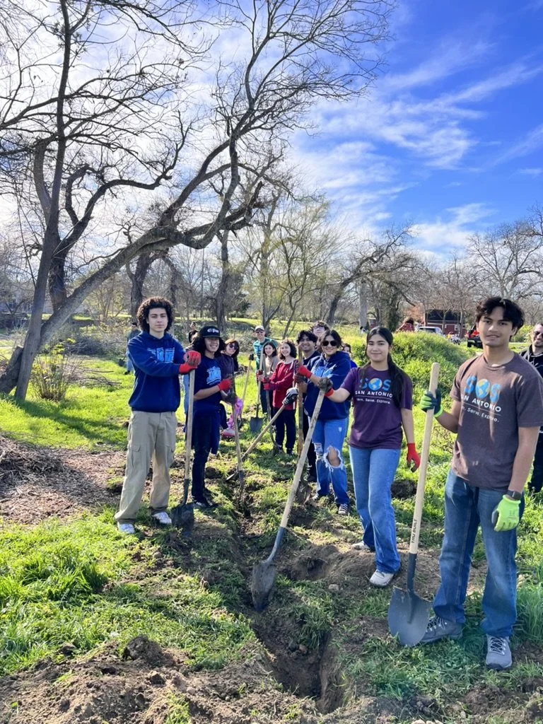 A group of young people participating in a tree planting activity outdoors on a sunny day, holding shovels, with some wearing gloves and SOS shirts standing in a line.