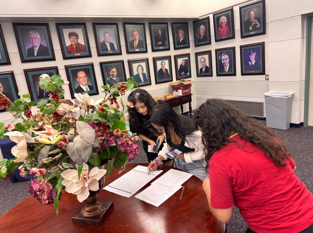 Three women gather around a wooden table with documents, in a room decorated with framed portraits on the wall and a large floral arrangement in the foreground.