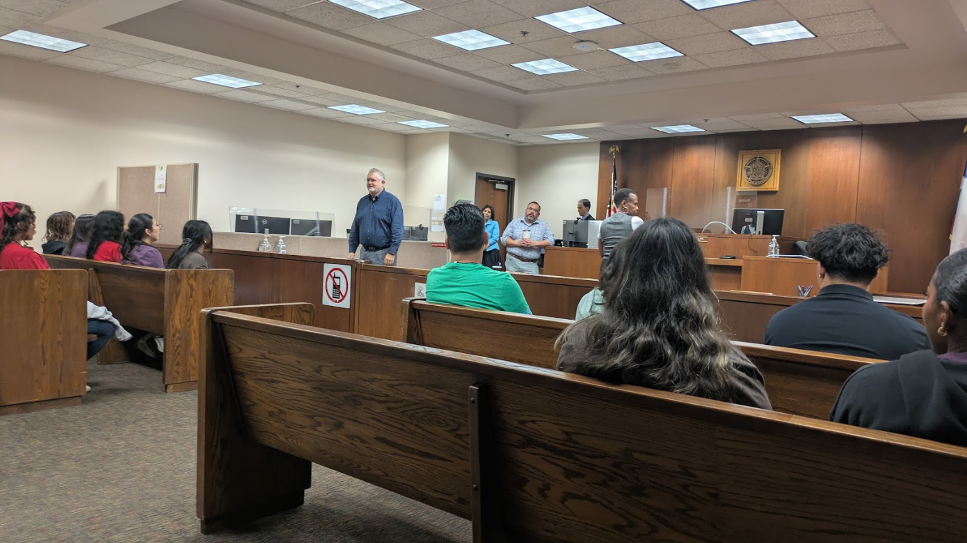 A courtroom with a judge's bench, where a judge and several other officials are seated. Several people are in the audience, some wearing red and purple clothing, and a man in a blue shirt stands in front of the judge's bench speaking. The room has wo