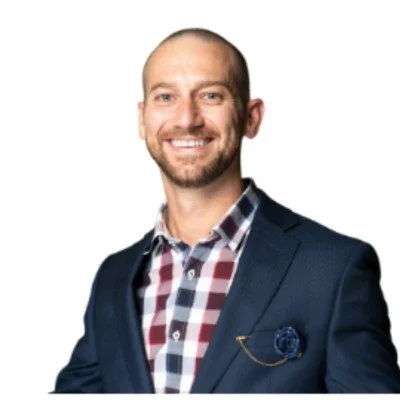 Smiling man in a checked shirt and navy blazer, with a lapel pin, against a white background.