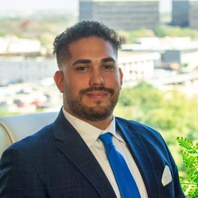 Professional man in a navy suit with blue tie and white pocket square, sitting in an office with cityscape view behind him.