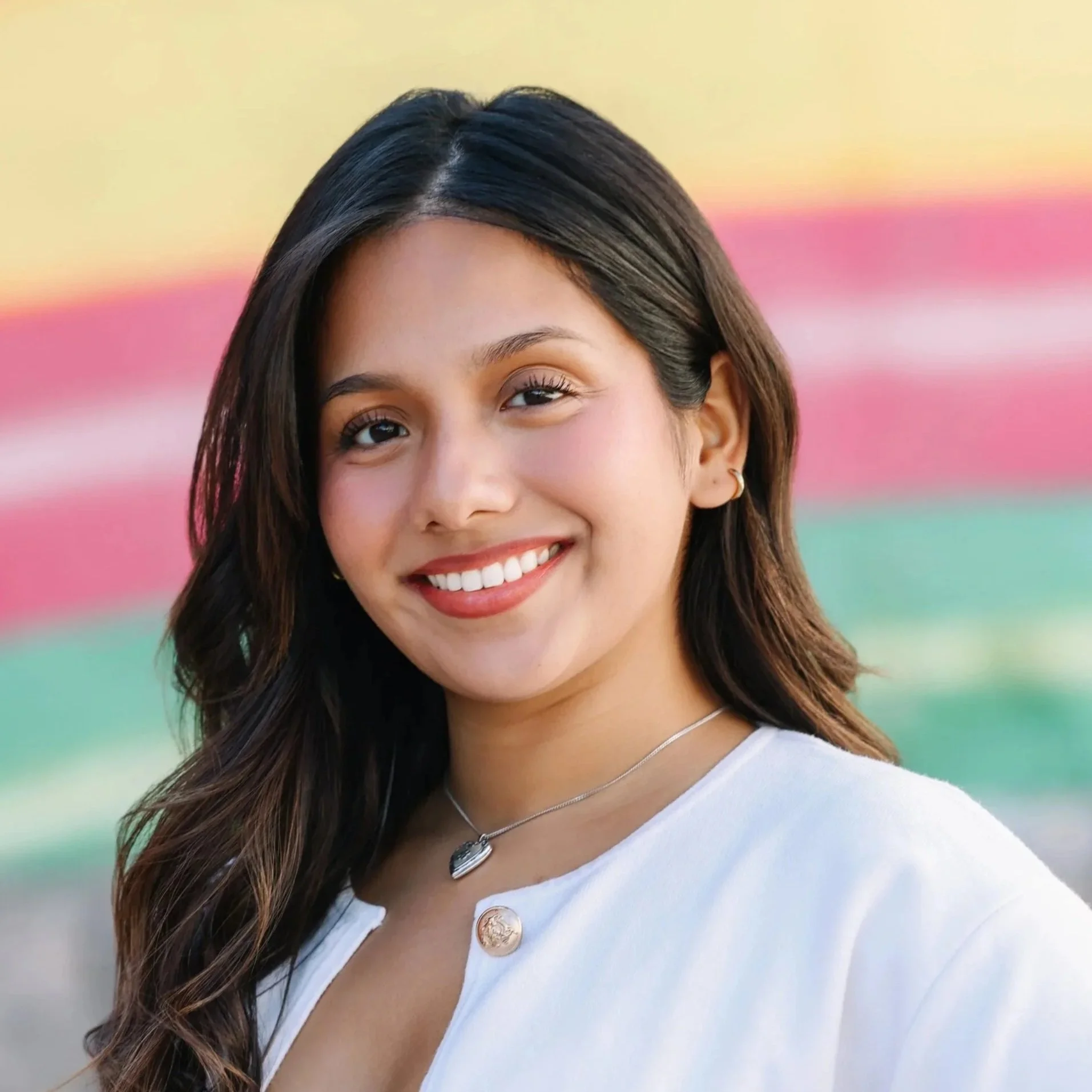 A young woman with long dark hair smiling outdoors with a colorful, blurred background of pink, yellow, and green.