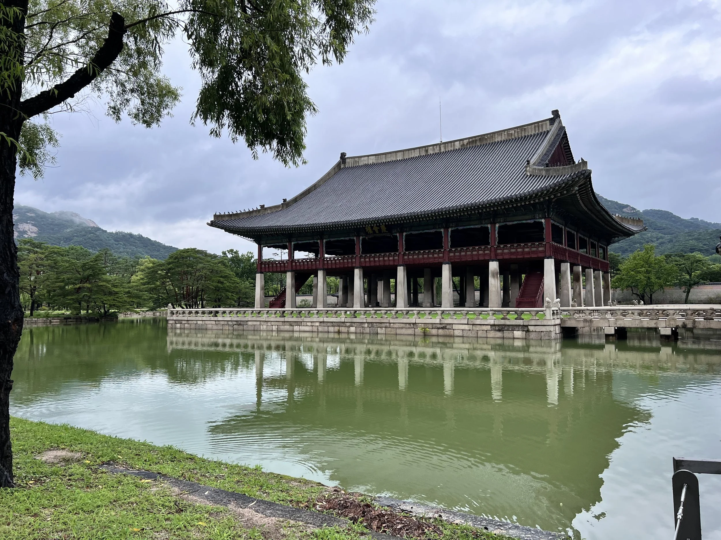 Traditional Asian pavilion with a tiled roof and red wooden accents, surrounded by a reflective pond, greenery, and mountains in the distance under a cloudy sky.