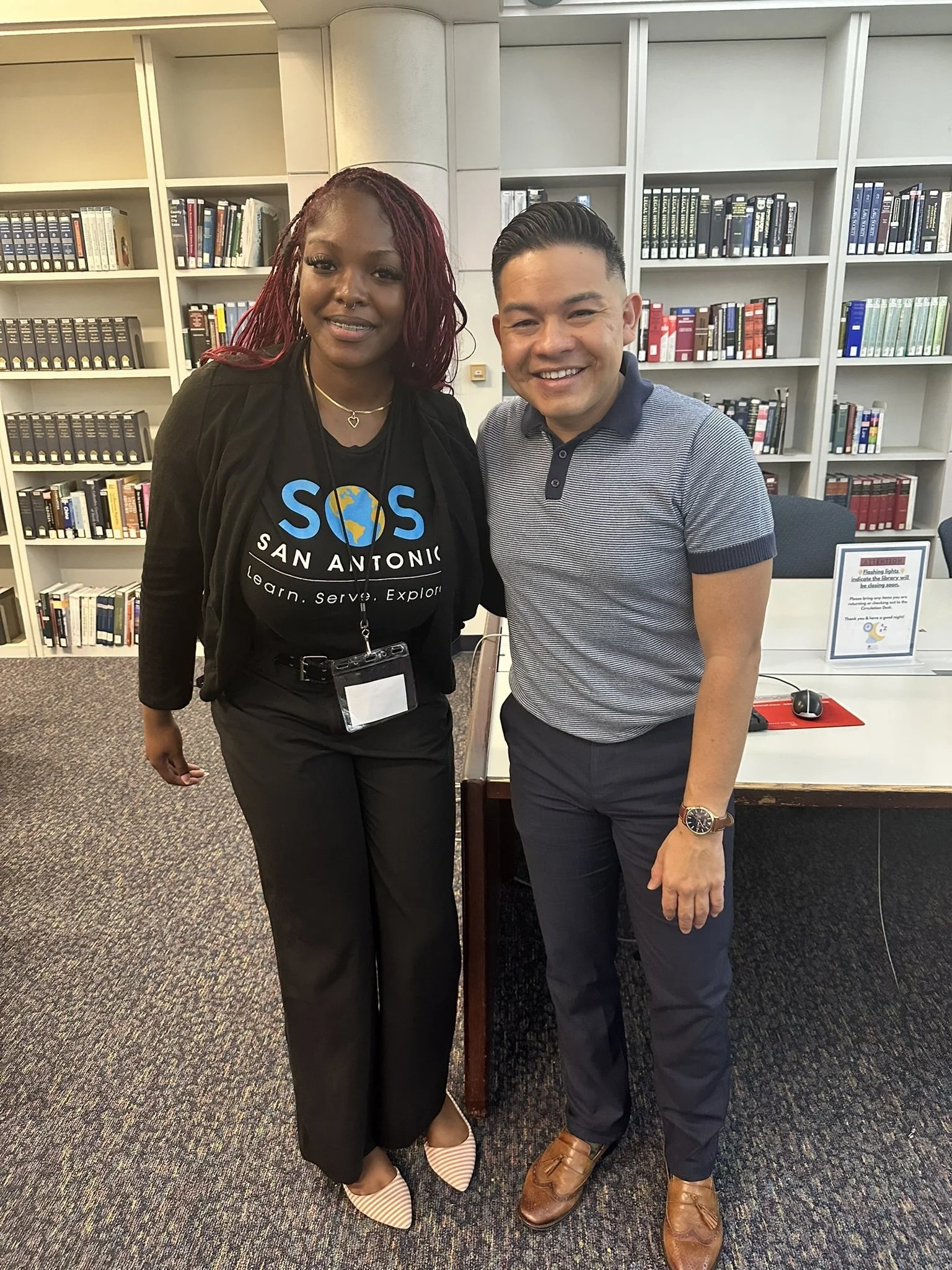 Two people standing side by side in a library, smiling at the camera. 