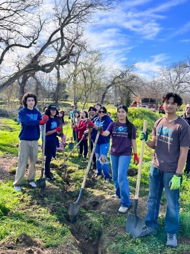 Group of young students planting trees outdoors on a bright sunny day, with some holding shovels and wearing gloves.