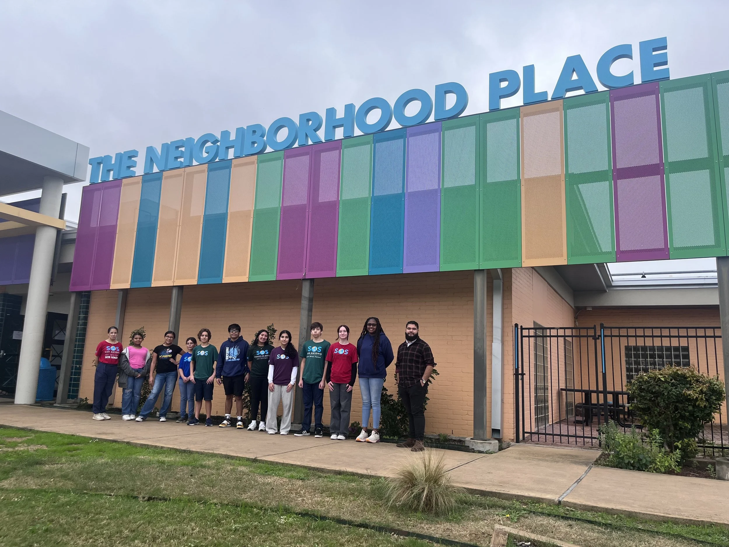 A group of people standing in front of a building with colorful panels and a sign that reads "The Neighborhood Place."