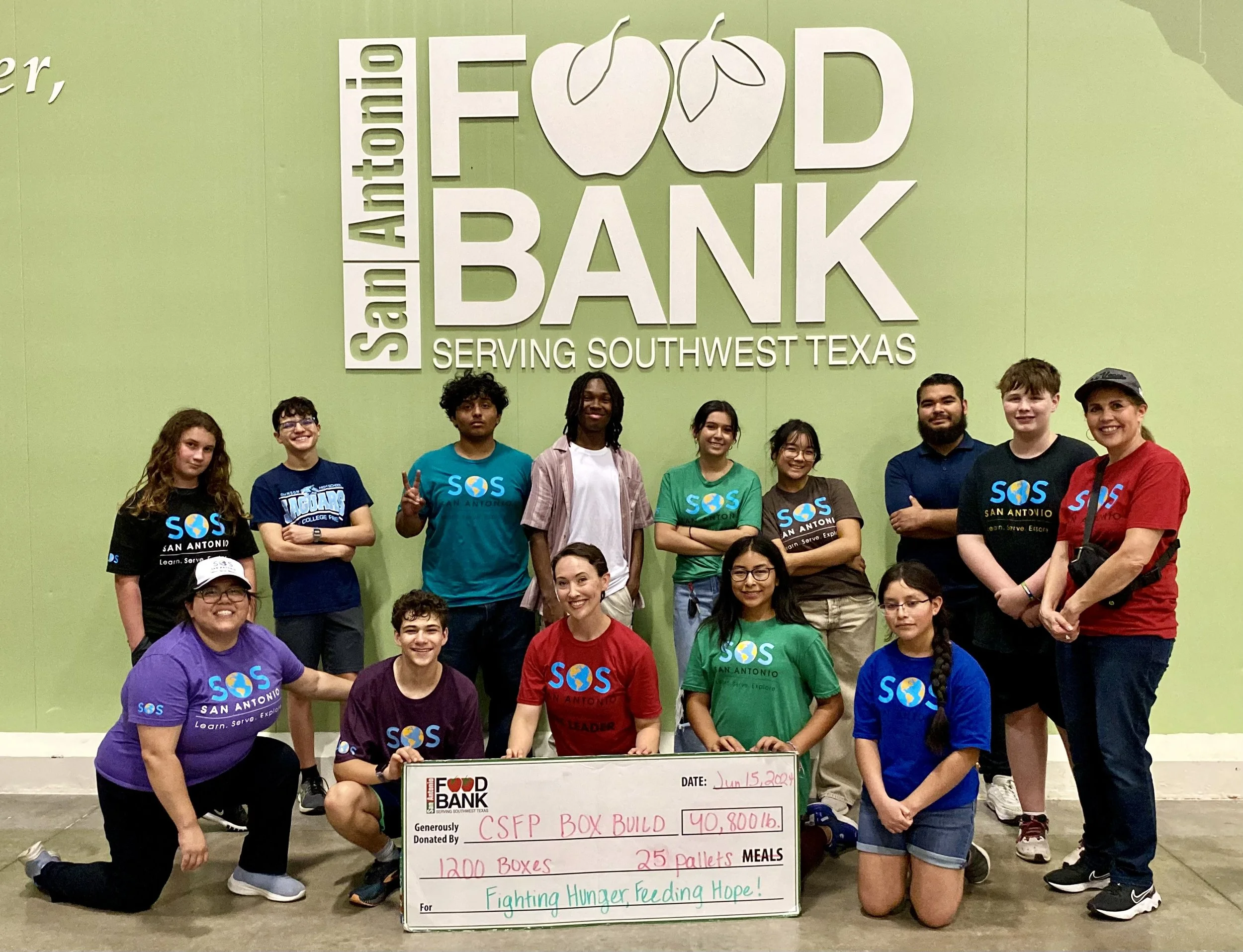 Group of young people and adults holding a large check at San Antonio Food Bank. The check shows donation details for 1200 boxes, 25 pallets, totaling $40,800, with a message 'Fighting Hunger, Feeding Hope'.