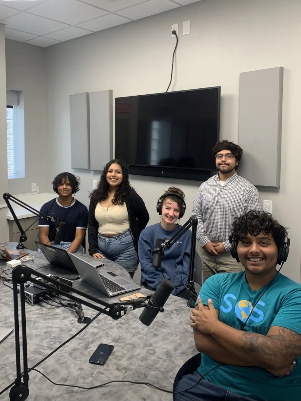 Five people in a recording studio posing for a photo, wearing headphones, with microphones, laptops, and recording equipment on a table.