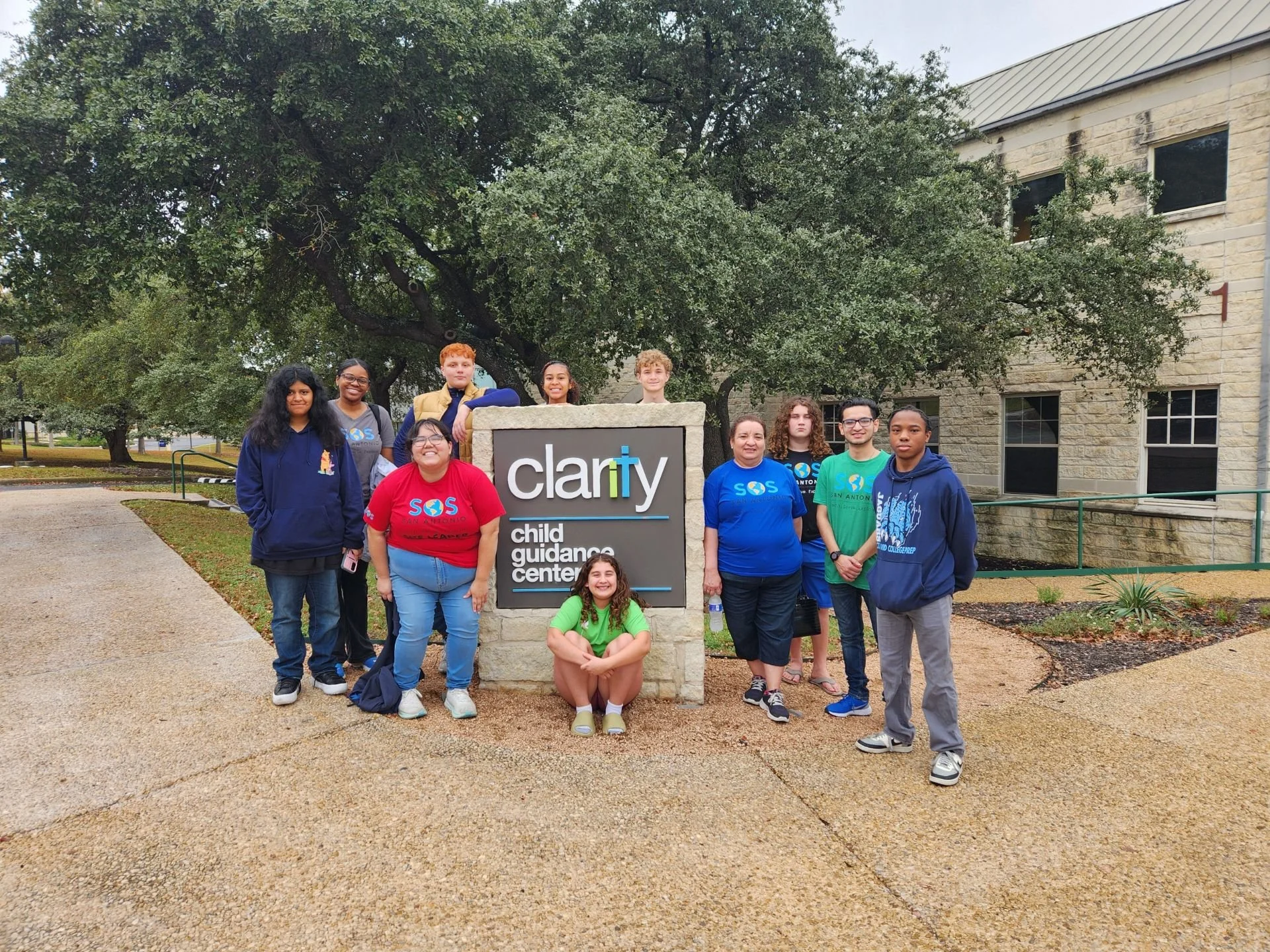 A group of ten diverse children and teenagers gathered around a sign that reads "clarity child guidance center" outside a brick building with a large tree in the background.