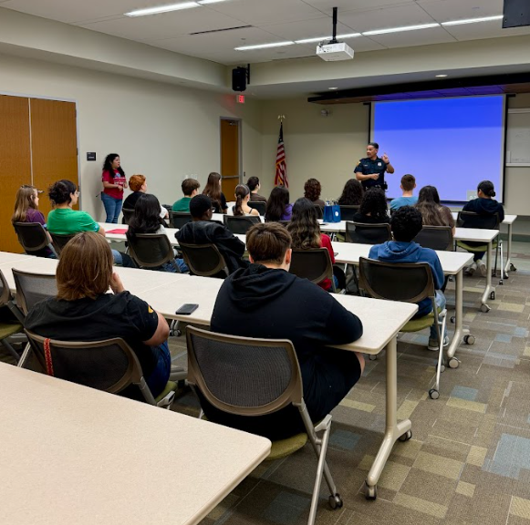 A group of students seated at desks in a classroom, listening to a man speaking at the front near a large screen and American flag.