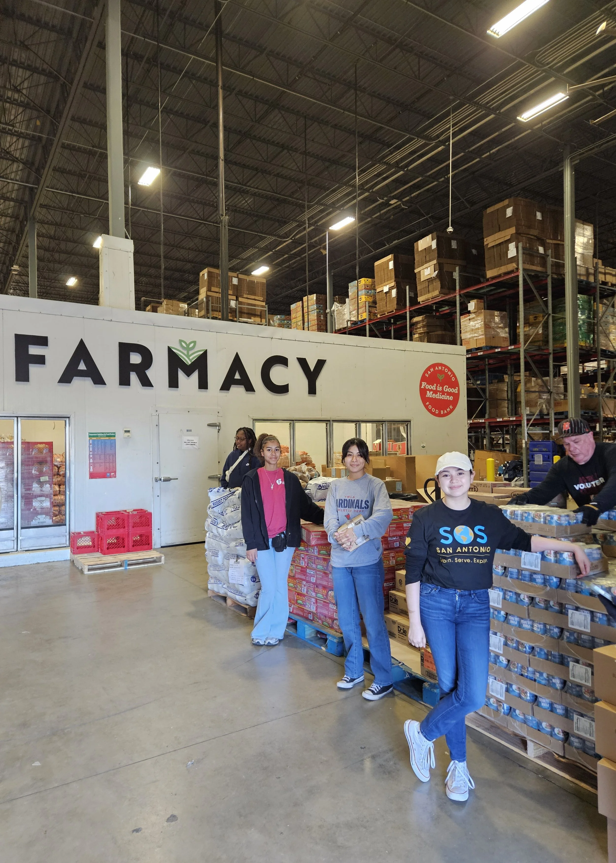 Four girls standing in front of a farmacy store inside a warehouse with shelves of food in the background. The girls are smiling and holding packages, with one volunteer wearing a black shirt with 'SOS San Antonio' printed on it.