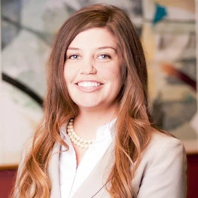 A woman with long, wavy, reddish-brown hair smiling at the camera, wearing a white blouse, a beige blazer, and a pearl necklace.