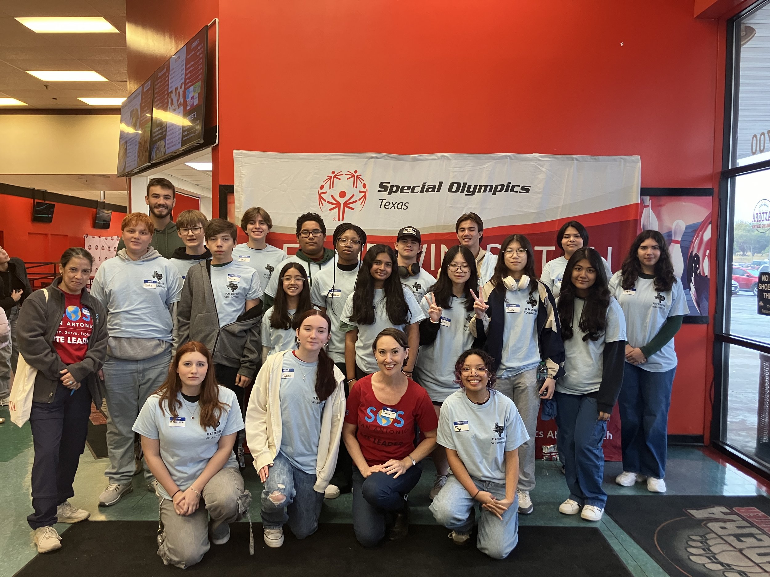 Group of young people standing in front of a banner that reads 'Special Olympics Texas' inside a building with a red wall and glass door.