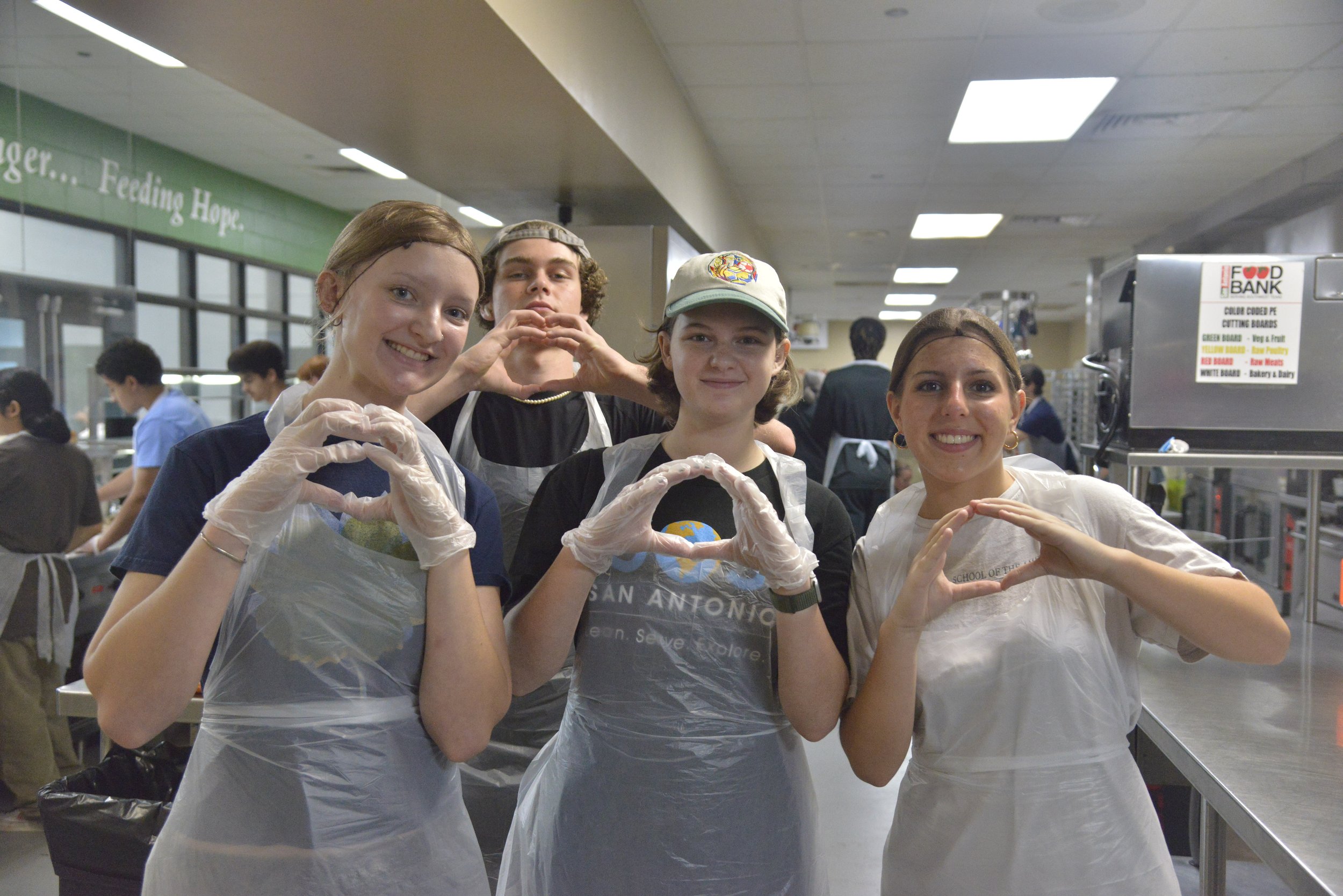 Four teenagers in a food bank, wearing gloves and aprons, forming heart shapes with their hands and smiling.