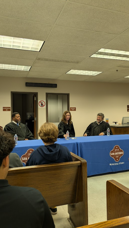Judicial panel in a courtroom with three judges seated at a table covered with a blue cloth displaying the San Antonio Municipal Court logo, and a person speaking at a podium.