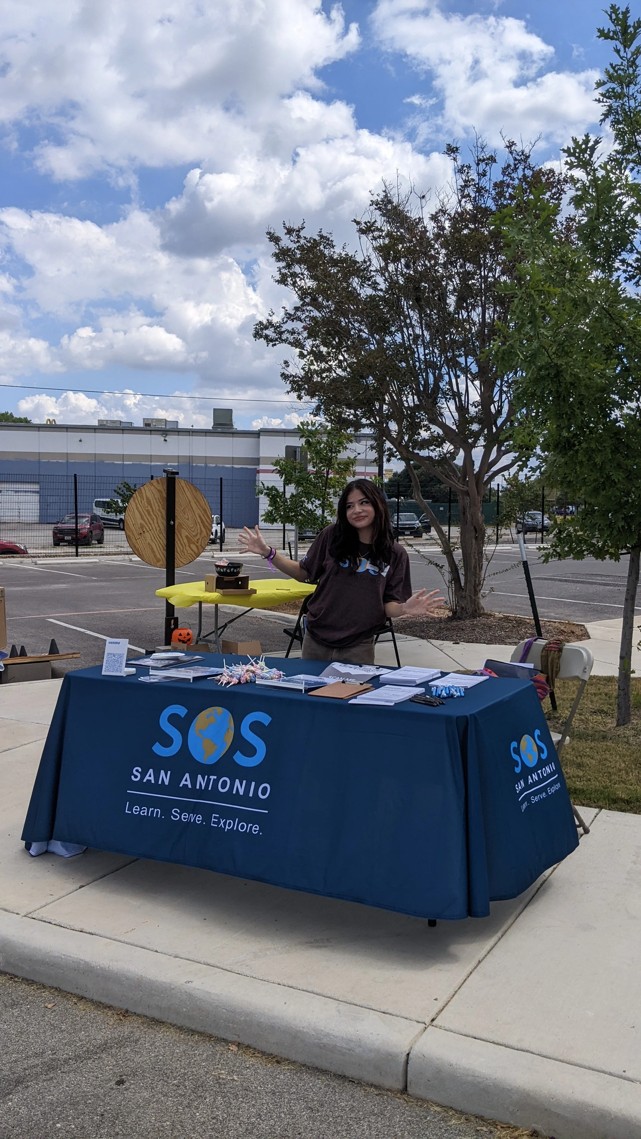 A woman standing behind a table with SOS San Antonio banner, outdoor setting with trees, parked cars, and a building in the background, during the day.