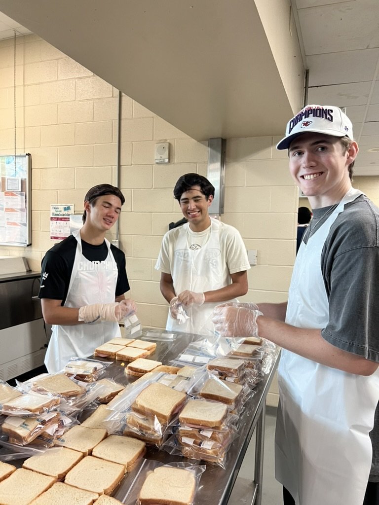 Three young men wearing aprons and gloves preparing sandwiches with sliced bread in a cafeteria or kitchen setting.